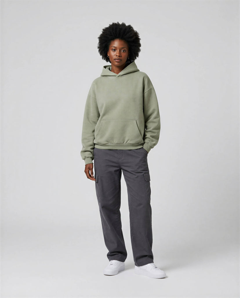 Blank heavyweight oversized hoodie in olive green, worn with gray cargo pants and white sneakers, standing against a neutr...